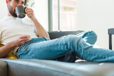 Genç sakallı adam elinde kahve fincanıyla kanepede dinleniyor.