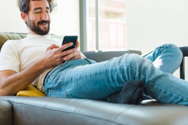 Genç sakallı adam akıllı bir telefonla kanepede dinleniyor.
