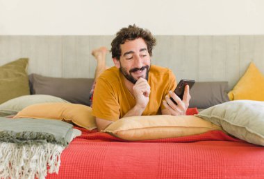 Yatakta cep telefonuyla sakallı genç bir adam.