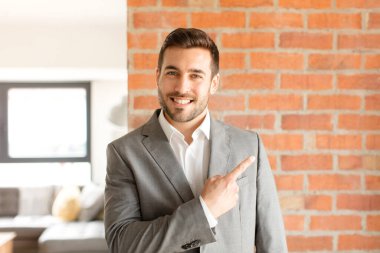 Yakışıklı iş adamı neşeli bir şekilde gülümsüyor, mutlu hissediyor ve kenarı ve yukarıyı işaret ediyor, fotokopi uzayındaki nesneleri gösteriyor.