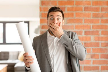 Yakışıklı mimar elleri ile ağzını kapatıyor, şok olmuş, şaşırmış bir ifadeyle, sır saklıyor ya da 