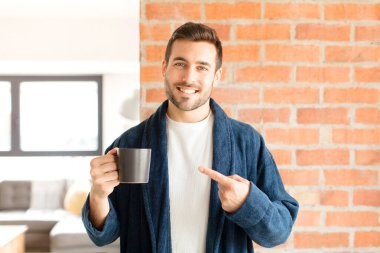 Yakışıklı adam neşeyle gülümsüyor, mutlu hissediyor ve yan ve yukarı doğru işaret ediyor, fotokopi uzayındaki nesneleri gösteriyor.