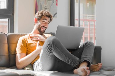 Genç sakallı adam komik bir şakaya gülüyor, mutlu ve neşeli hissediyor, dizüstü bilgisayarla kanepe konseptinde eğleniyor.