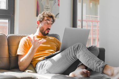 Genç sakallı adam, ağzı açık ve şaşırmış, şok olmuş ve hayrete düşmüş bir şekilde dizüstü bilgisayarla kanepe konseptinde inanılmaz bir sürpriz yapmış.