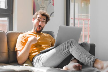 Genç sakallı adam şok olmuş ve şaşırmış görünüyor. Ağzını açmış, dizüstü bilgisayarla kanepe konseptini işaret ediyor.