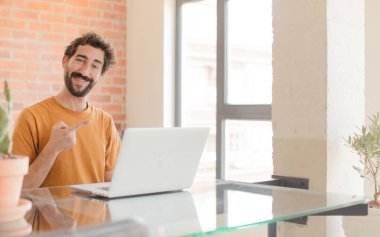 Genç sakallı adam neşeyle gülümsüyor, mutlu hissediyor ve yan ve yukarı doğru işaret ediyor, fotokopi uzayındaki nesneleri gösteriyor ve bir dizüstü bilgisayarla çalışıyor.