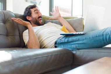 Genç sakallı bir adam dizüstü bilgisayarla kanepede dinleniyor.