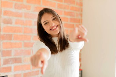 Genç bir kadın mutlu ve kendinden emin, kamerayı iki eliyle gösteriyor ve gülüyor, seni seçiyor.