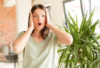 Güzel bir kadın hoş olmayan bir şekilde şok olmuş, korkmuş ya da endişeli, ağzı sonuna kadar açık ve iki kulağını da eliyle kapatıyor.