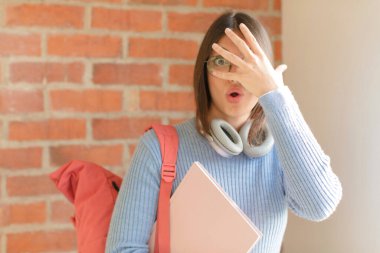 Güzel bir öğrenci şok olmuş, korkmuş ya da korkmuş görünüyor. Yüzü elle örtülmüş ve parmaklarının arasından gizlice bakıyor.