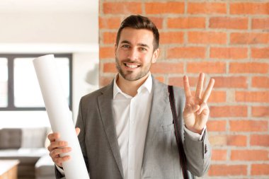 Yakışıklı mimar gülümsüyor ve arkadaş canlısı görünüyor. Üç ya da üç numarayı gösteriyor. Elleri önde, geri sayıyor.