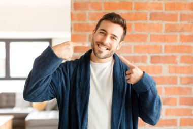 Yakışıklı adam kendinden emin bir şekilde gülümsüyor. Geniş bir gülümseme, pozitif, rahat, tatmin olmuş bir tavır sergiliyor.