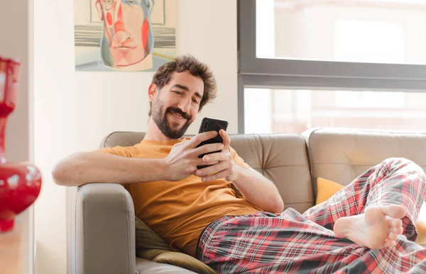 Genç sakallı adam akıllı bir telefonla kanepede oturuyor.