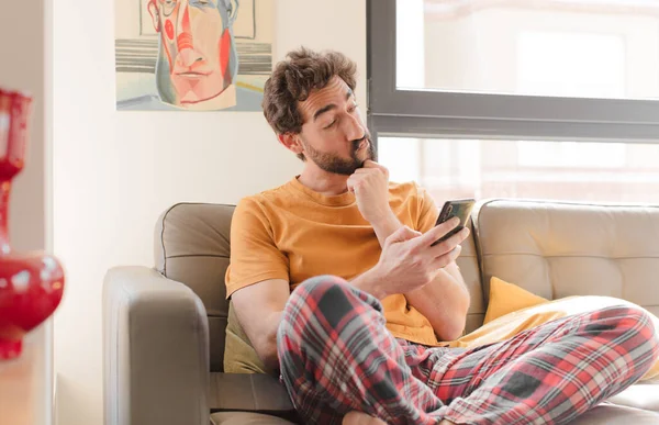 Genç sakallı adam akıllı bir telefonla kanepede oturuyor.