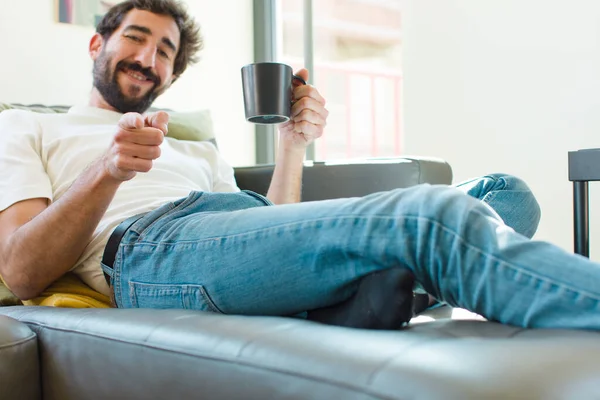 若い髭を生やした男がコーヒーマグカップを持ってソファに腰を下ろして — ストック写真