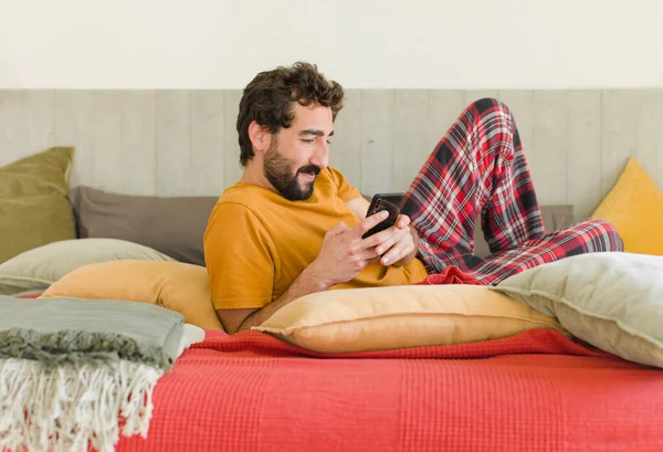 Yatakta cep telefonuyla sakallı genç bir adam.