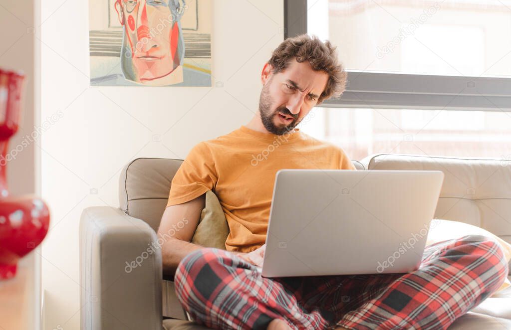 young bearded man feeling puzzled and confused, with a dumb, stunned expression looking at something unexpected and sitting with a laptop