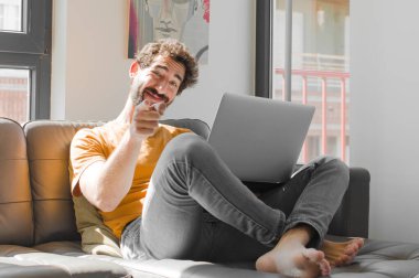 Genç sakallı adam kameraya tatmin olmuş, kendinden emin, arkadaş canlısı bir gülümsemeyle bakıyor seni dizüstü bilgisayarla kanepe konseptinde seçiyor.