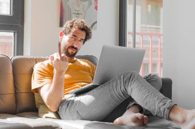 Genç sakallı adam sinirli, sinirli, asi ve agresif hissediyor, orta parmağını şıklatıyor, dizüstü bilgisayarla kanepe konseptinde savaşıyor.