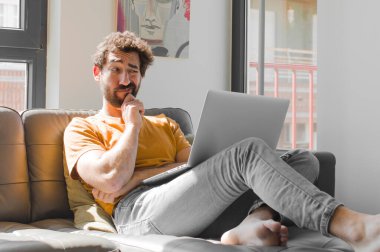 Genç sakallı adam düşünüyor, kuşkulu ve şaşkın hissediyor, farklı seçeneklerle, bir dizüstü bilgisayarla hangi kararı vereceğini merak ediyor.