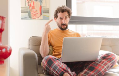 Genç sakallı adam şaşkın ve şaşkın hissediyor, deli olduğunu ya da aklını kaçırdığını ve dizüstü bilgisayarla oturduğunu gösteriyor.