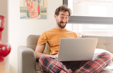 Genç sakallı adam mutlu ve hoş bir şekilde şaşırmış görünüyor, büyülenmiş ve şok olmuş bir ifadeyle heyecanlı ve dizüstü bilgisayarla oturuyor.