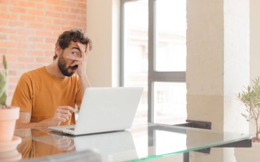 Genç sakallı adam şok olmuş, korkmuş ya da dehşete düşmüş görünüyor. Yüzü elle örtülmüş, parmaklarının arasından gizlice bakıyor ve bir dizüstü bilgisayarla çalışıyor.