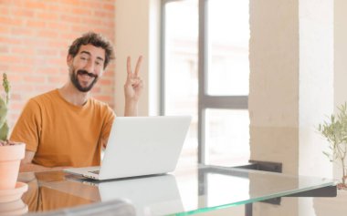 Genç sakallı adam gülümsüyor ve arkadaş canlısı görünüyor. İki ya da iki numara önde, geriye doğru sayıyor ve dizüstü bilgisayarla çalışıyor.