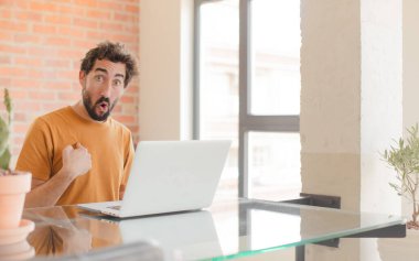 Genç sakallı adam ağzı açık, şok olmuş ve şaşırmış görünüyor. Kendini işaret ediyor ve bir dizüstü bilgisayarla çalışıyor.