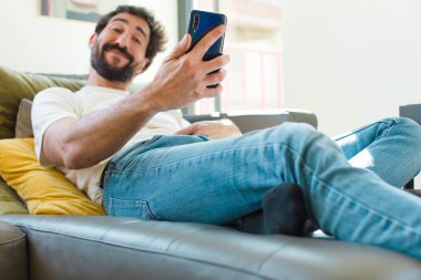 Genç sakallı adam akıllı bir telefonla kanepede dinleniyor.