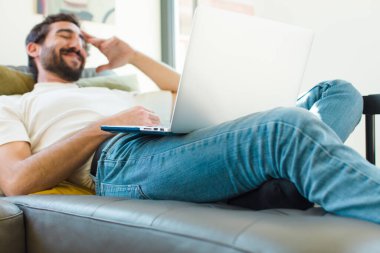 Genç sakallı bir adam dizüstü bilgisayarla kanepede dinleniyor.