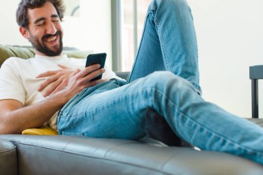 Genç sakallı adam akıllı bir telefonla kanepede dinleniyor.