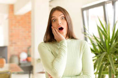 Güzel bir kadın şok ve inançsızlık içinde, elleri yanağında ve kolları çaprazlanmış, sersemlemiş ve şaşırmış hissediyor.