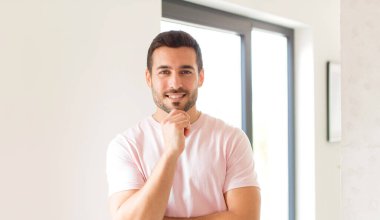 Yakışıklı adam mutlu görünüyor ve gülümsüyor. Elleri çenesinde, merak ediyor ya da soru soruyor. Seçenekleri karşılaştırıyor.