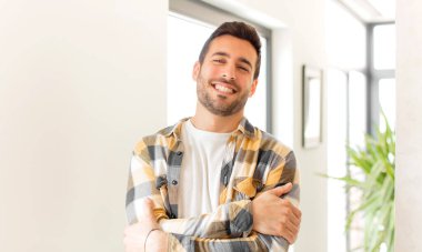 Yakışıklı adam kollarını kavuşturup mutlu bir şekilde gülüyor, rahat, pozitif ve tatmin edici bir pozla.