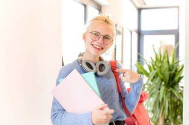 Güzel öğrenci neşeli bir şekilde gülümsüyor, mutlu hissediyor ve kenarı ve yukarıyı işaret ediyor, kopyalama alanında nesneleri gösteriyor.