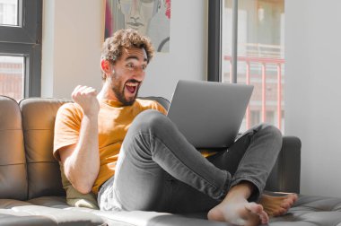 Genç sakallı adam şok olmuş, heyecanlı ve mutlu hissediyor, gülüyor ve başarıyı kutluyor, vay canına! Dizüstü bilgisayarlı kanepe konsepti.