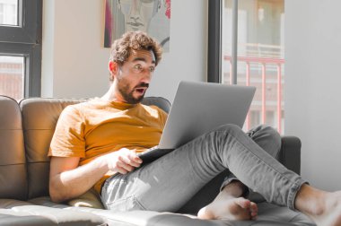 Genç sakallı adam şok olmuş ya da şaşırmış görünüyor, ağzı açık bir şekilde dizüstü bilgisayarla kanepe konseptinde 