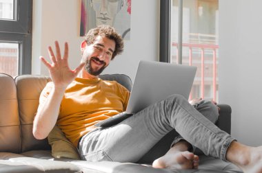 Genç sakallı adam gülümsüyor ve arkadaş canlısı görünüyor. 5 ya da 5 numarayı gösteriyor. Elinde dizüstü bilgisayarla kanepe konseptinde geriye doğru sayıyor.