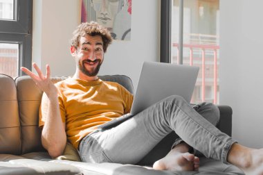 Genç sakallı adam mutlu, şaşırmış ve neşeli hissediyor, olumlu bir tavırla gülümsüyor, bir çözüm ya da fikri bir dizüstü bilgisayarla kanepe konseptinde fark ediyor.