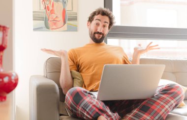 Genç sakallı adam şaşkın ve şaşkın hissediyor, şüpheci, farklı seçenekleri tartıyor ya da seçiyor komik bir ifadeyle ve dizüstü bilgisayarla oturuyor