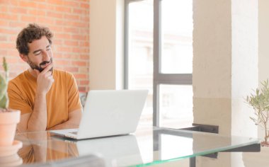 Genç sakallı adam mutlu, kendinden emin bir ifadeyle gülümsüyor. Elleri çenesinde, merak ediyor, yan tarafa bakıyor ve bir dizüstü bilgisayarla çalışıyor.
