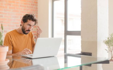 Genç sakallı adam şaşkın ve şaşkın hissediyor, deli olduğunu ya da aklını kaçırdığını ve bir dizüstü bilgisayarla çalıştığını gösteriyor.