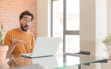 Genç sakallı adam şaşırmış ve şok olmuş görünüyor, çenesi düşmüş, eli açık bir objeyi tutuyor ve bir dizüstü bilgisayarla çalışıyor.