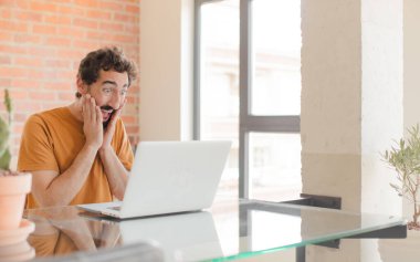 Genç sakallı adam mutlu, heyecanlı ve şaşırmış hissediyor. İki eli yüzü üzerinde ve dizüstü bilgisayarla çalışıyor.