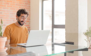 Genç sakallı adam mutlu ve hoş bir şekilde şaşırmış görünüyor. Büyülenmiş ve şok olmuş bir ifadeyle heyecanlı ve bir dizüstü bilgisayarla çalışıyor.