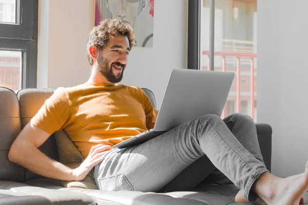 Genç sakallı adam mutlu bir şekilde gülümsüyor, eli kalçasında ve kendinden emin, pozitif, gururlu ve arkadaş canlısı bir tavır içinde, dizüstü bilgisayarında bir dizüstü bilgisayarla.