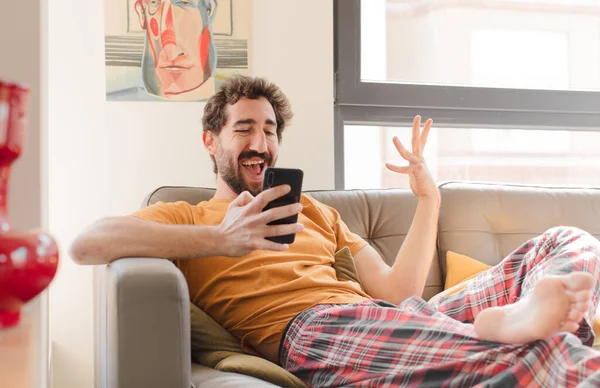 Genç sakallı adam akıllı bir telefonla kanepede oturuyor.