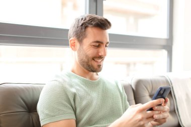 Genç yakışıklı adam akıllı telefonunu evde kullanıyor.