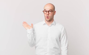 Kel adam şaşırmış ve şoka uğramış görünüyor, çenesi düşmüş ve eli açık bir nesneyi tutuyor.